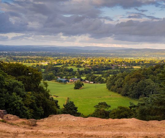 A scenic view of Alderley Edge's countryside with green fields, scattered houses, clusters of trees, and a cloudy sky in the background.
