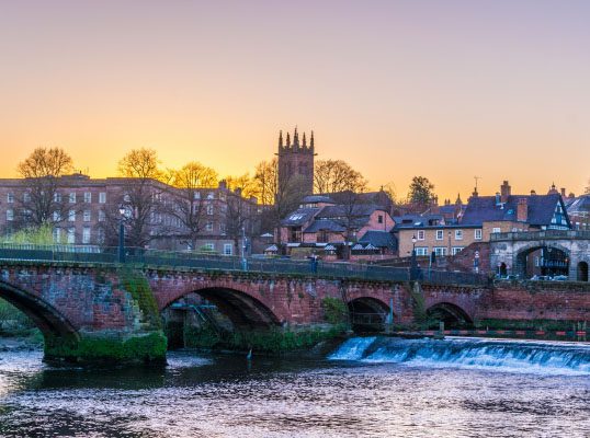 The picture shows a scenic view of a historic stone bridge over a flowing river with buildings and a church tower in the background at sunset, reminiscent of the charming landscapes in Chester.