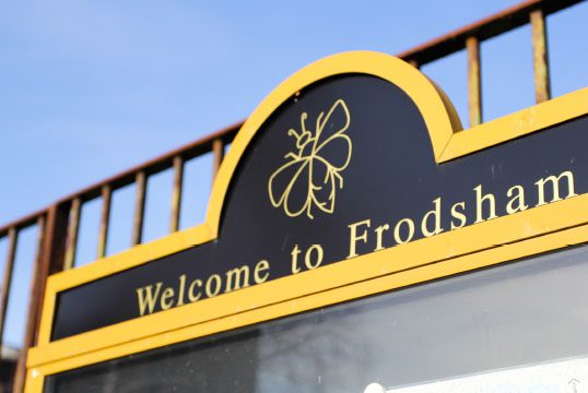 Sign with a yellow border reading "Welcome to Frodsham," featuring a butterfly silhouette above the text. The background shows part of a railing and a clear blue sky.
