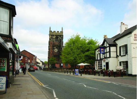 A street scene in Holmes Chapel shows a church with a clock tower, flanked by buildings on either side. A few cars are parked on the left and tables with umbrellas are set up outside a building on the right.