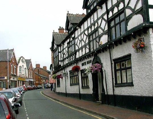 A street in Middlewich with parked cars, featuring a historic black-and-white timber-framed building adorned with hanging flower baskets and additional buildings further down the street.