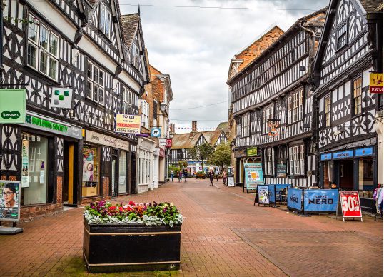 A narrow pedestrian street in Nantwich is lined with black and white Tudor-style buildings. A flowerbed sits in the foreground, and various shops and signs are visible along the street, adding a charming touch to this picturesque scene.