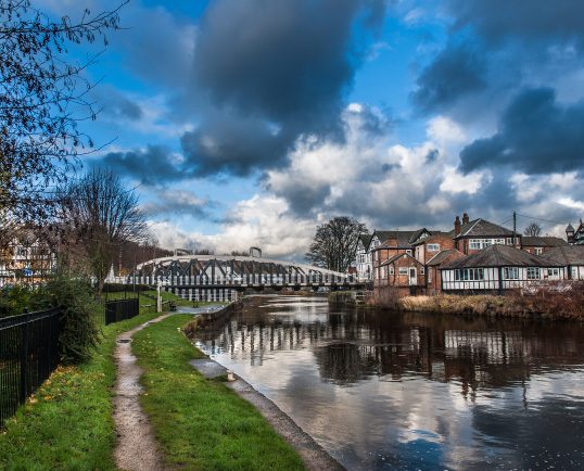 A path runs alongside a tranquil river with houses and a bridge in the background under a partly cloudy sky, reminiscent of the serene landscapes in Northwich.