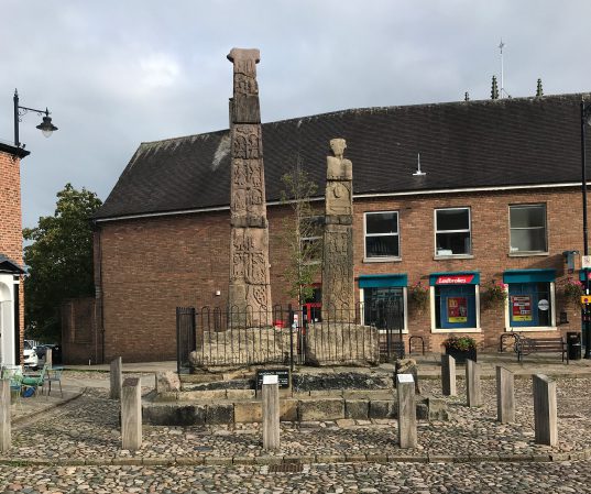 Two tall, intricately carved stone pillars stand in a cobblestone square in Sandbach, enclosed by a low fence and surrounded by modern brick buildings and a few shops.