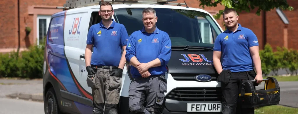 Three men wearing blue shirts and work pants stand in front of a white JBL Julian Bland Heating and Plumbing company van and a ladder on top, ready for their next home project.