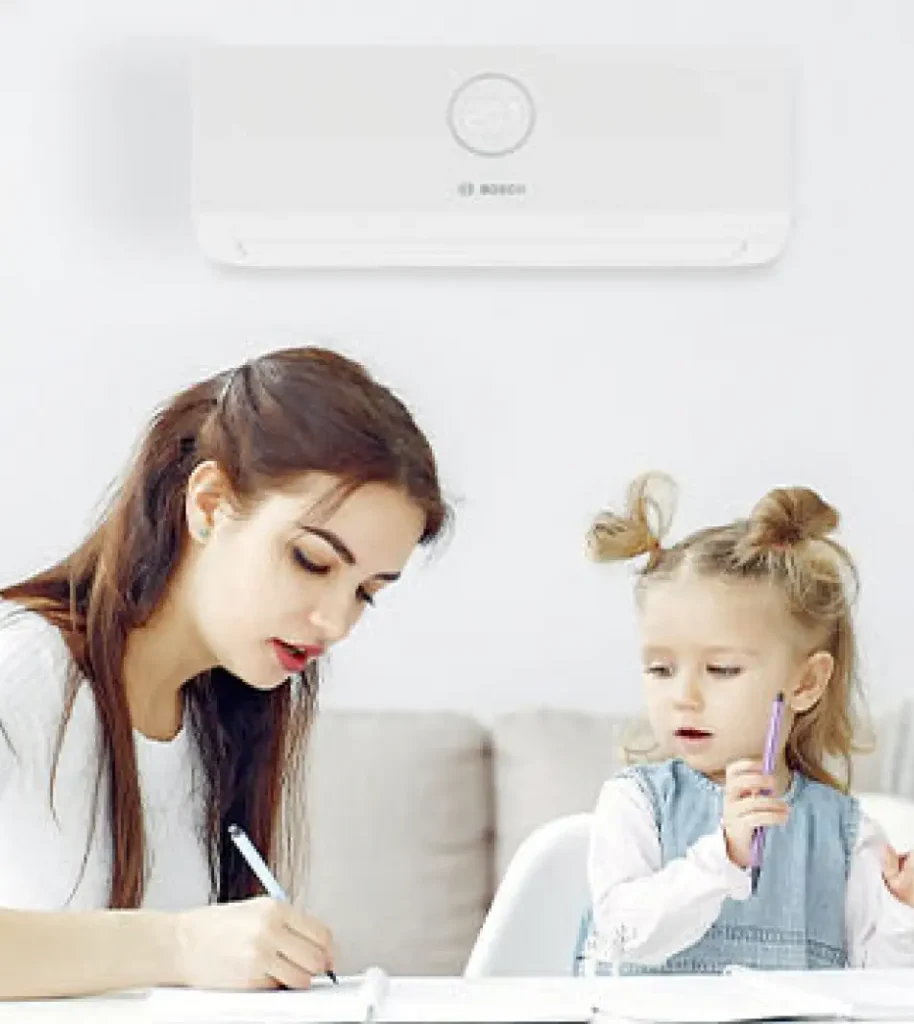 A woman and a child sit at a table working on activities in the comfort of their home. An air conditioner is mounted on the wall above them.