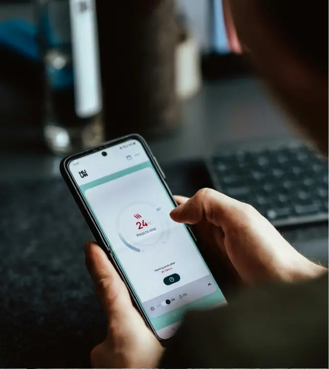 A person holding a smartphone with a temperature monitoring app showing 24 degress on the screen. Bespoke Heating Systems brochures are visible next to the keyboard in the background.