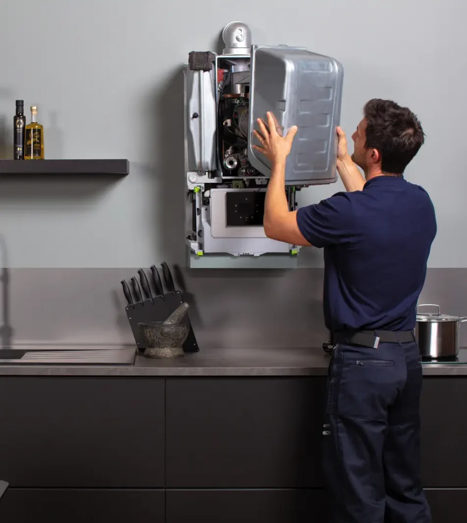 A technician in a blue uniform works on a mounted kitchen appliance, perhaps a boiler or water heater, with kitchen items like knives and pots in the background, ensuring the home heating system is functioning efficiently.