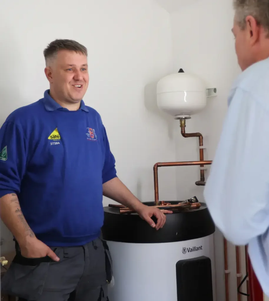 A man in a blue work uniform stands next to a white water heater, conversing with another man in the comfort of a home.