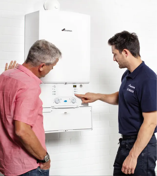 Two men standing near a wall-mounted boiler. The man in a navy blue shirt is explaining the control panel to the man in a red shirt, emphasizing key points of proper boiler servicing.