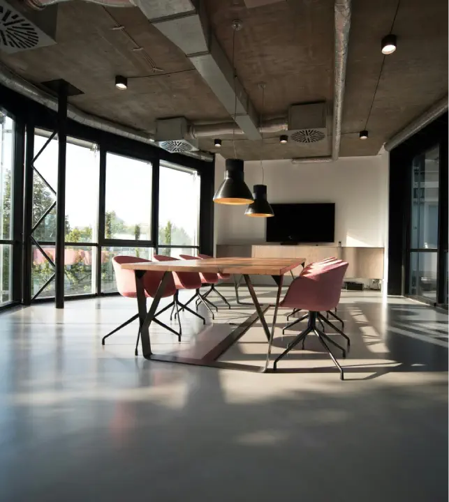 A modern, commercial conference room with a long wooden table, pink chairs, large windows, heating systems, and industrial-style ceiling fixtures.