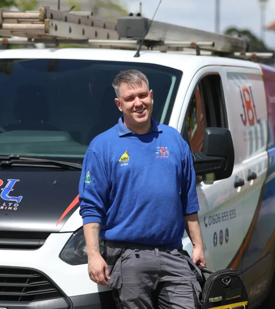 Danny Bland wearing a blue long-sleeve shirt stands next to JBL company van with ladders on its roof and company logos on the side. With short hair and carrying a tool bag, he represents the professionalism and dedication.