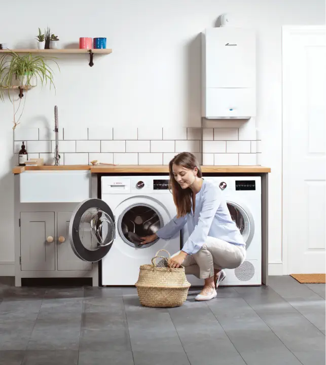 A woman in a laundry room loads clothes into a front-loading washing machine. There are plants on a shelf and a laundry basket on the floor, next to a sleek Worcester boiler. The room also has a white tiled backsplash and cabinets, adding to its neat appearance.