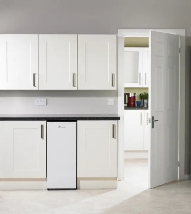 A minimalist kitchen with white cabinets, a compact refrigerator below the countertop, and an open door leading to a pantry with additional storage, appliances, and an efficient oil boiler.