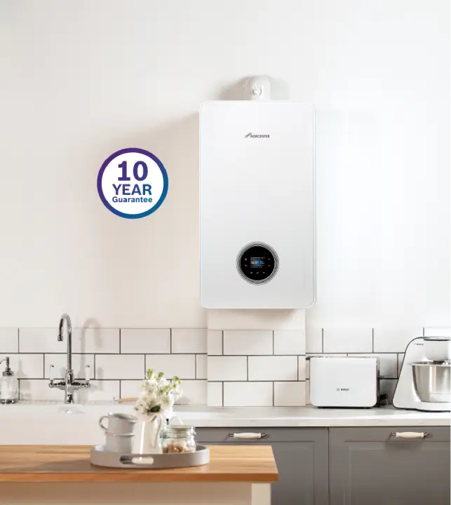 A white boiler, part of an advanced home heating system, with a digital display is mounted on a kitchen wall above the counter. To the side, there is a "10 Year Guarantee" sign. The kitchen counter features a pot, toaster, and stand mixer.
