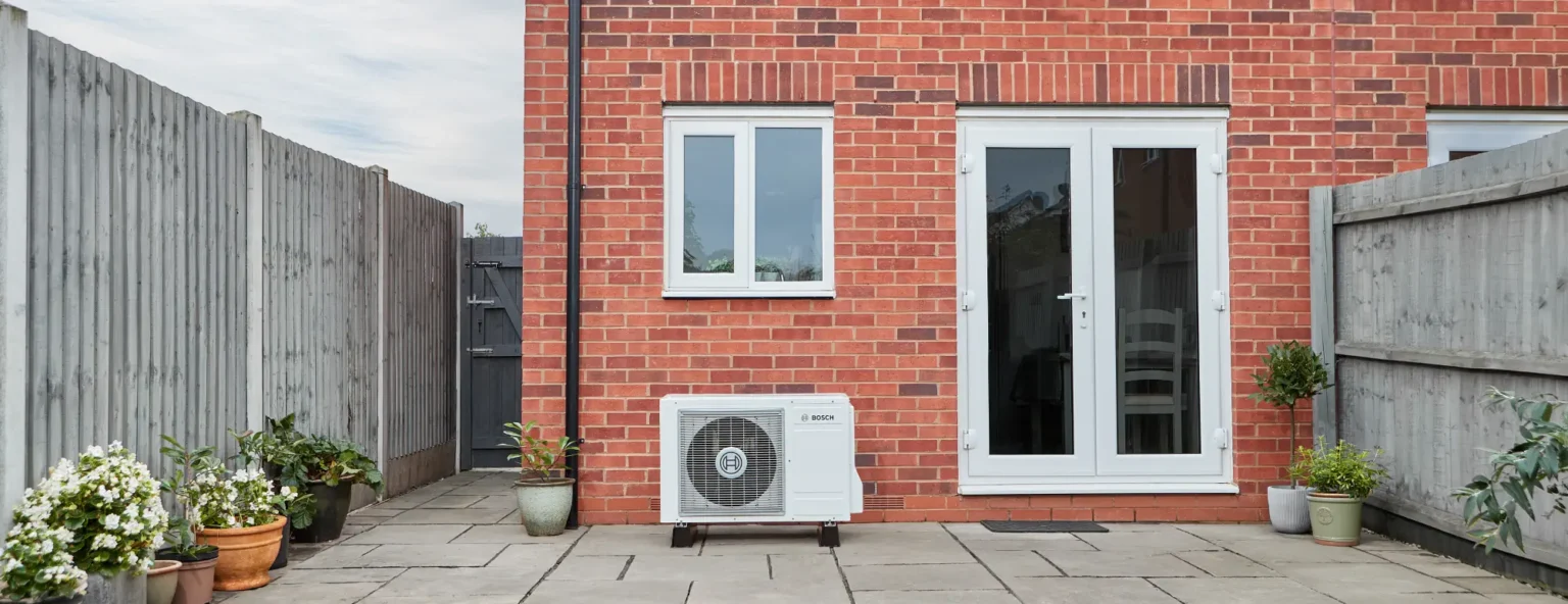 A red brick house with white-framed windows and white double glass doors leading to a paved patio. An outdoor air conditioning unit, part of an energy-efficient heat pump system, is installed on the patio. Green grass in the foreground.