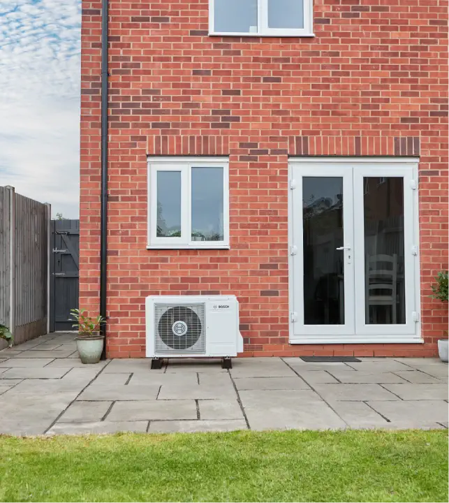 A red brick house with white-framed windows and white double glass doors leading to a paved patio. An outdoor air conditioning unit, part of an energy-efficient heat pump system, is installed on the patio. Green grass in the foreground.