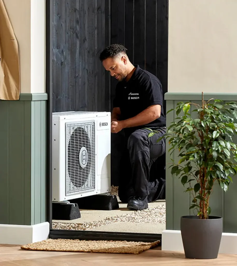 A technician kneels while working on an outdoor heat pump unit, ensuring everything runs smoothly for the home. A potted plant is visible in the foreground inside the house.