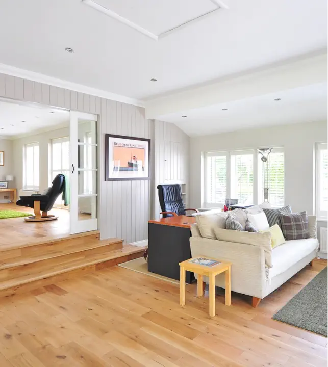 A bright living room with wooden floors, a white sofa, a black armchair, and large windows letting in natural light. An underfloor heating system keeps the area cozy. Steps lead to another comfortable space with an additional armchair and a rug.