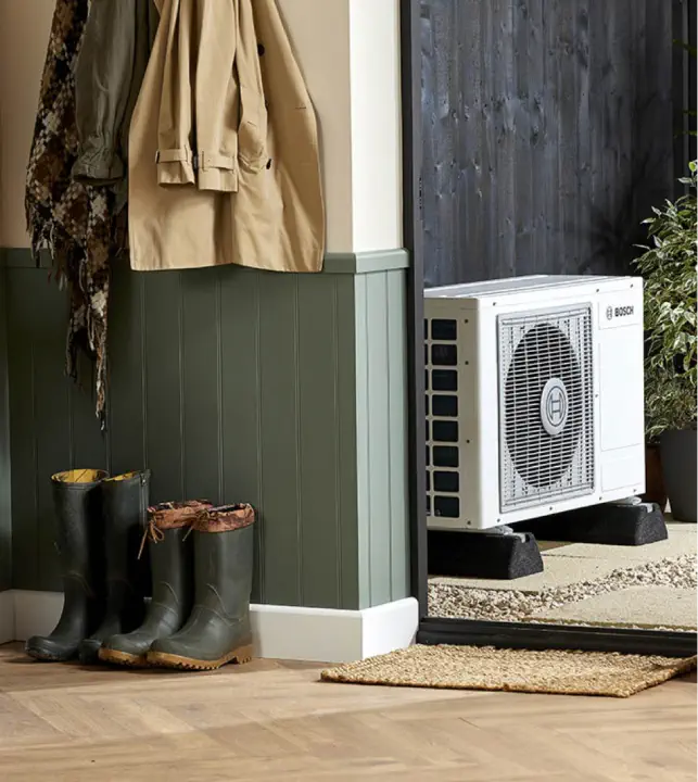 Coats hang on wall pegs next to a pair of boots on the floor; a bespoke air conditioning unit marked "Bosch" is positioned outside near a door.