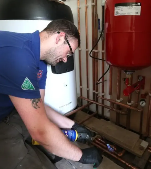 A person wearing a blue shirt and black gloves uses a handheld tool to work on copper pipes in the mechanical room, tending to boiler maintenance. Red and white machinery are visible in the background.