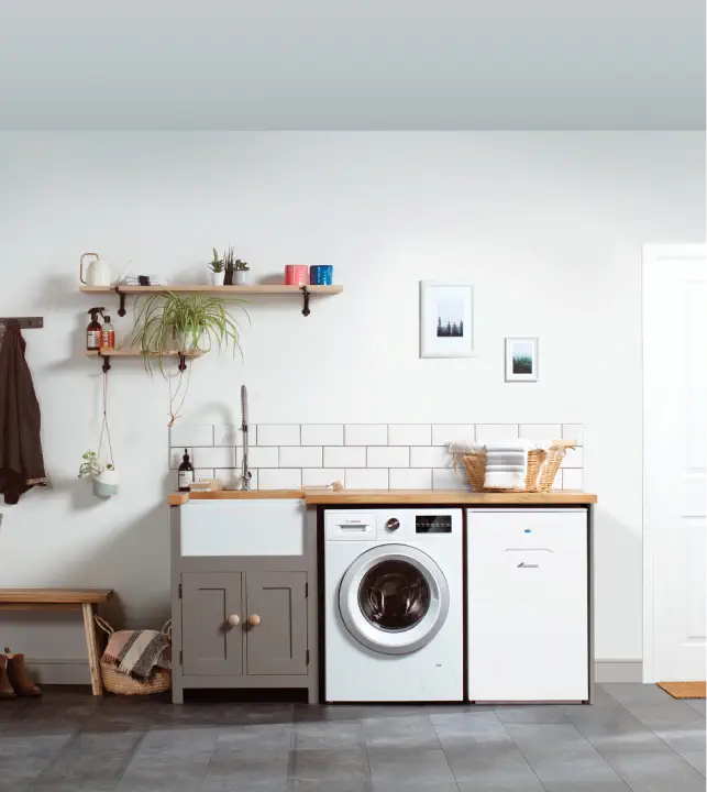 A laundry room with a washing machine, a dryer, an oil boiler discreetly tucked in the corner, a sink with a wooden countertop, a shelf holding plants and toiletries, a coat rack, and a small bench against a white tiled wall.
