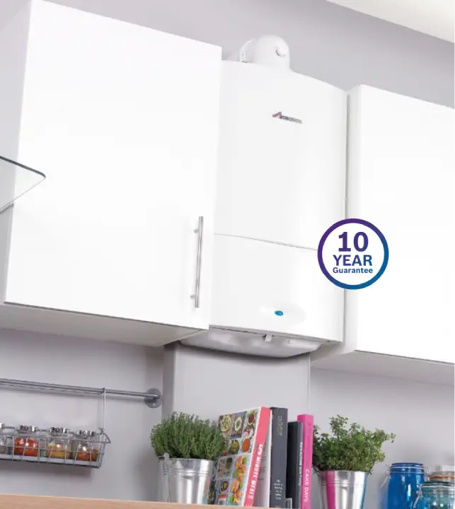 White kitchen cabinets with a mounted white boiler featuring a "10 Year Guarantee" sticker. This modern heating system is complemented by a shelf below, holding potted herbs and cookbooks.