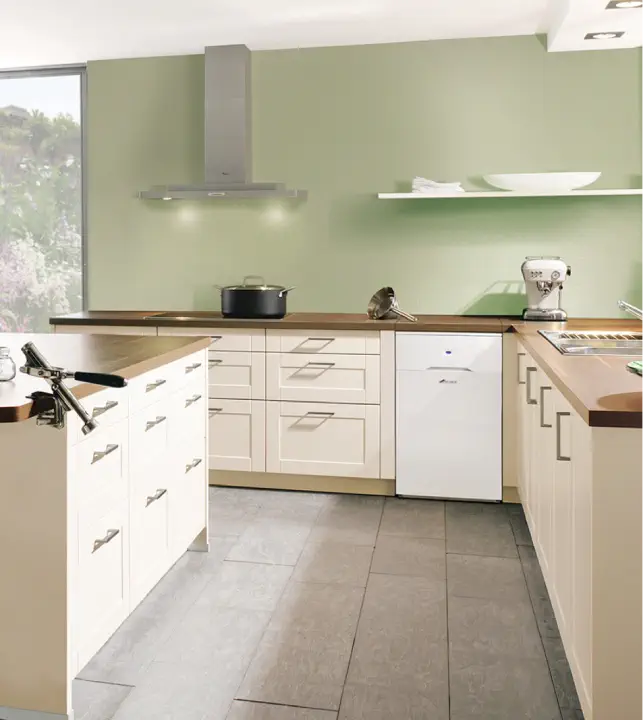 Modern kitchen with light green walls, a central island with drawers, and white cabinetry. Stainless steel range hood, white dishwasher, countertop appliances, and discreetly tucked-away oil boilers add functional elegance to the space.