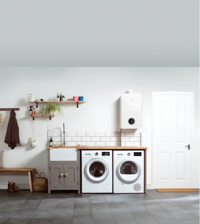 A laundry room with a wooden bench, a shelf, a coat rack, a counter with a sink, and neatly tucked in the corner are the washing machine, dryer, and modern gas boilers.