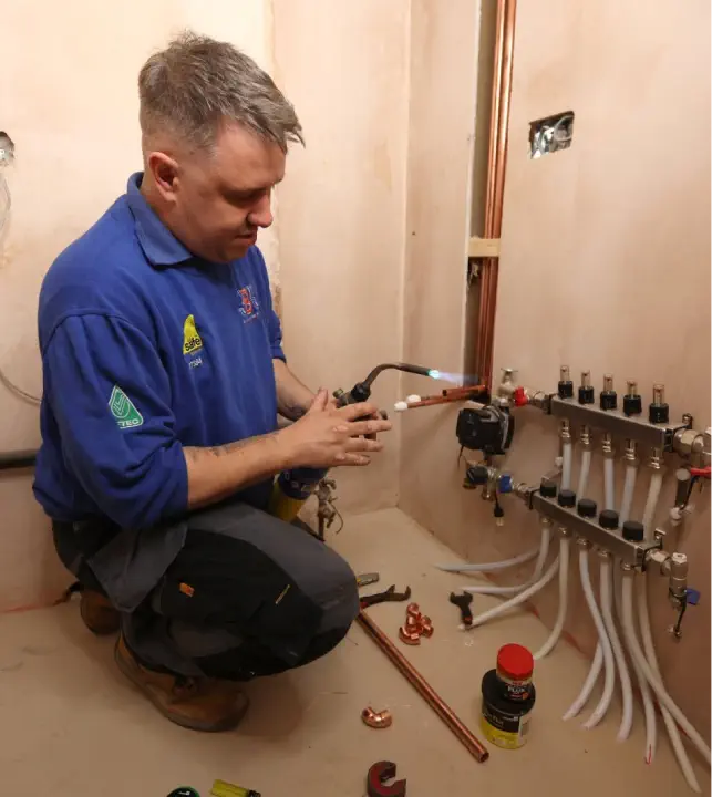 A man in a blue shirt is kneeling and using a blowtorch to work on copper pipes connected to gas boilers in a room with various plumbing components. Tools and materials are scattered around him.