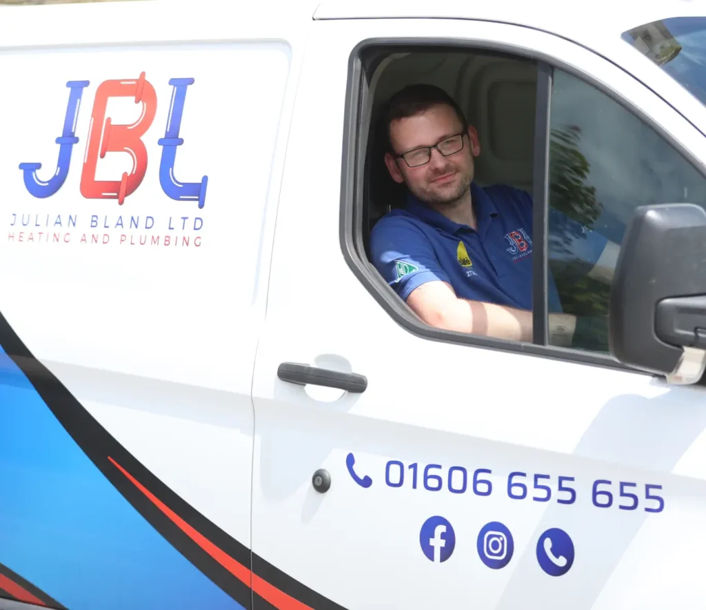 A JBL technician wearing glasses and a blue shirt is sitting inside a white van marked with Julian Bland Ltd Heating and Plumbing. The van, essential for home repairs, displays contact information and social media icons.